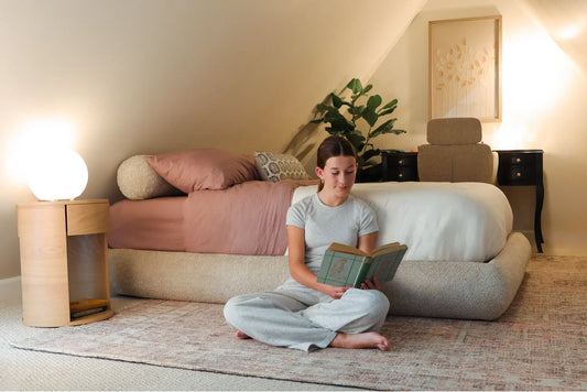 Girl sitting on the floor reading a book beside a cozy upholstered SoftFrame designs bed frame with soft lighting and neutral decor.