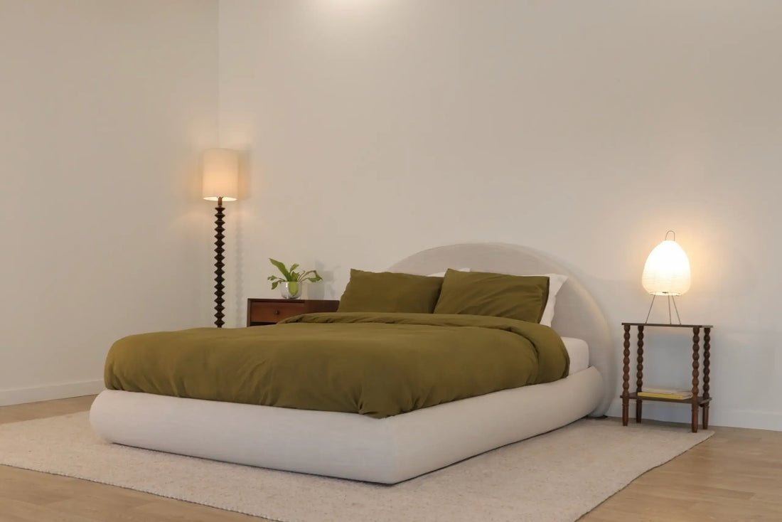 A minimalist bedroom with a low, upholstered SoftFrame Designs bed featuring olive green bedding and pillows. The bed sits on a light-colored rug atop a wooden floor.