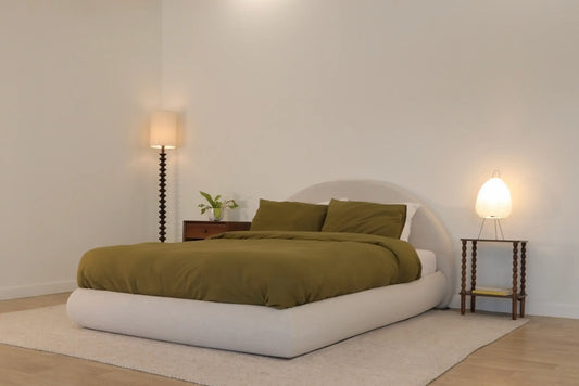 A minimalist bedroom with a low, upholstered SoftFrame Designs bed featuring olive green bedding and pillows. The bed sits on a light-colored rug atop a wooden floor.