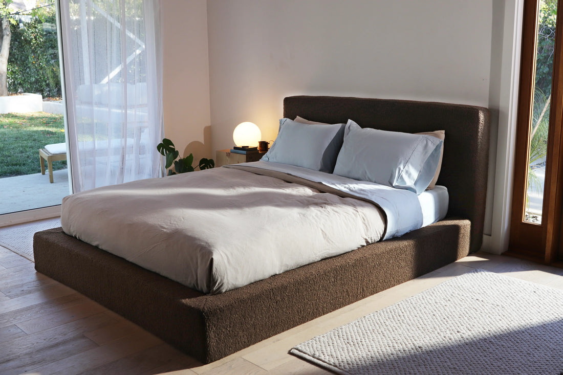 Sunlit minimalist bedroom with an upholstered SoftFrame Designs bed, neutral bedding, and a small bedside lamp by large glass doors overlooking a garden