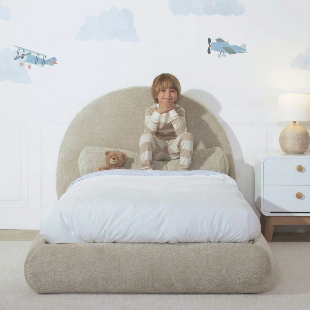 Child sitting on a large bed with teddy bear, airplane wall decals, and nightstand.