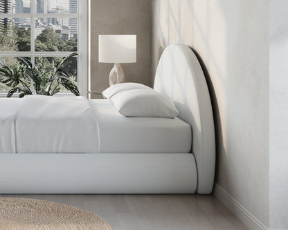 Modern bedroom with a curved all cushioned headboard, lamp, and window view of trees.