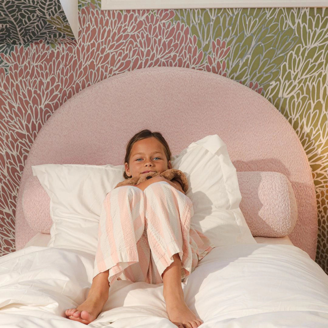 Child sitting on a bed with a pink all cushioned headboard and floral wallpaper.