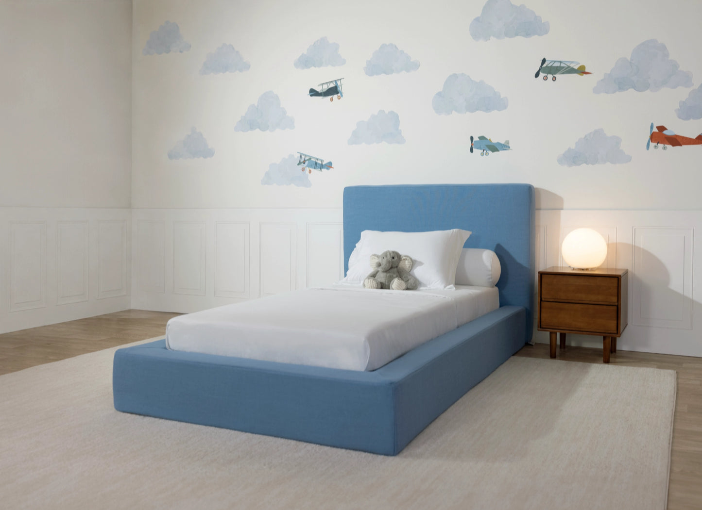 A modern platform bed with a light blue upholstered frame and headboard, styled with white bedding and a plush elephant pillow, set in a child's bedroom with cloud and airplane wall decals.