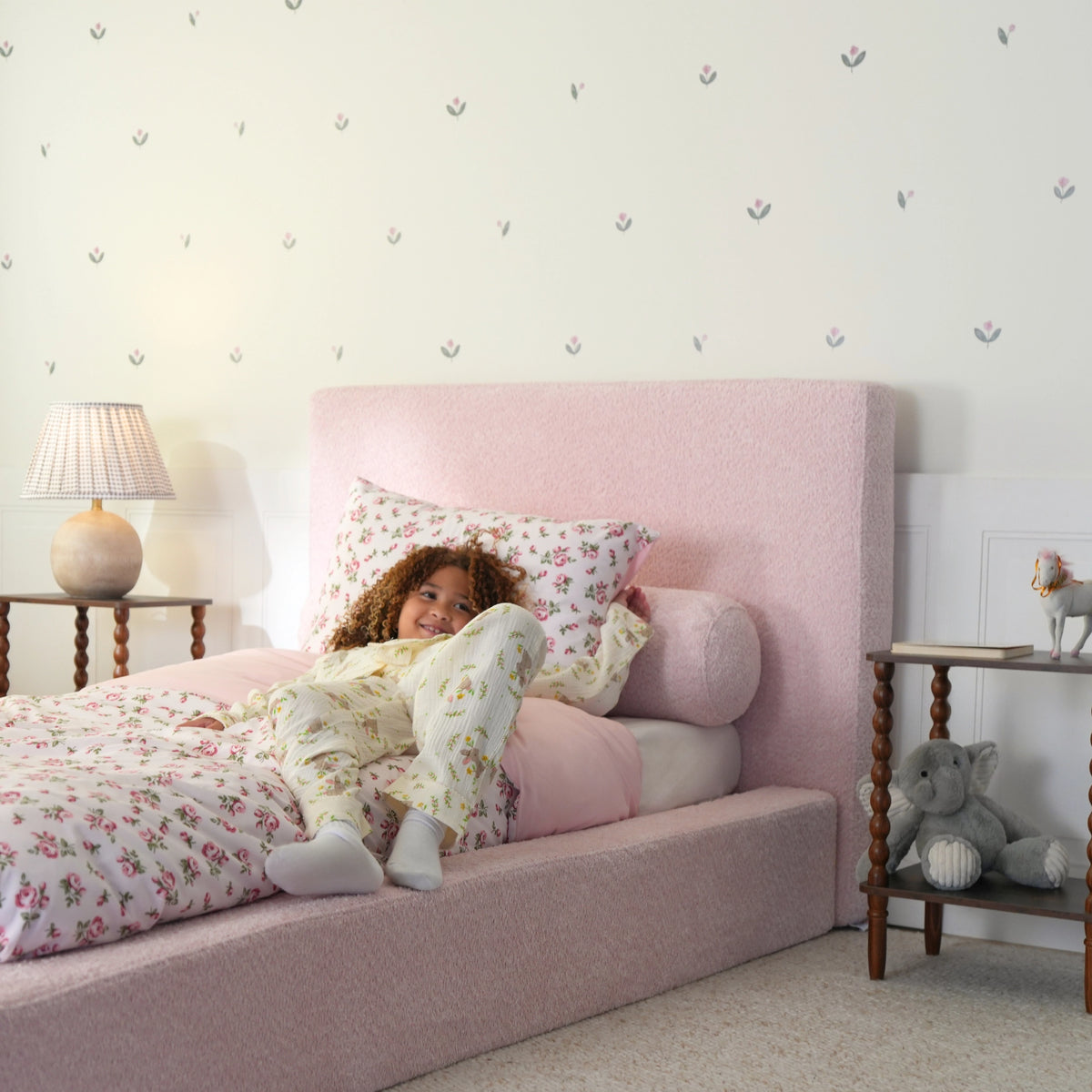 side view image of a girl sitting on a pink all cushioned bed bed with floral bedding.