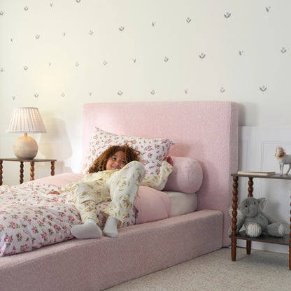 side view image of a girl sitting on a pink all cushioned bed bed with floral bedding.
