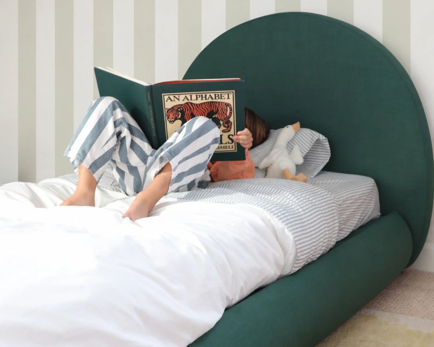 boy reading a book in bed with a green all cushioned headboard and striped wallpaper.