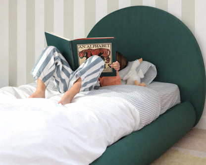 boy reading a book in bed with a green all cushioned headboard and striped wallpaper.