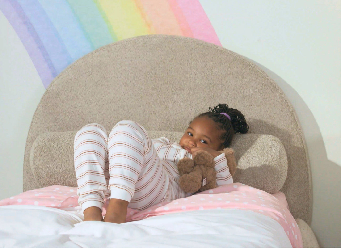 A young child wearing pink and white striped pajamas lies on a bed with a pink polka-dot blanket, holding a brown teddy bear. The background features a large beige curved all-cushioned headboard and a pastel rainbow painted on the wall.