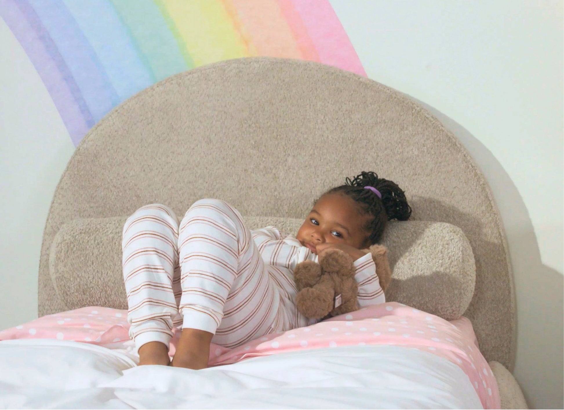 A young child wearing pink and white striped pajamas lies on a bed with a pink polka-dot blanket, holding a brown teddy bear. The background features a large beige curved all-cushioned headboard and a pastel rainbow painted on the wall.