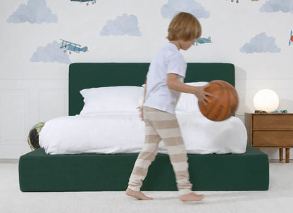 Child playing with a basketball in a bedroom with green all cushioned bed and white walls.