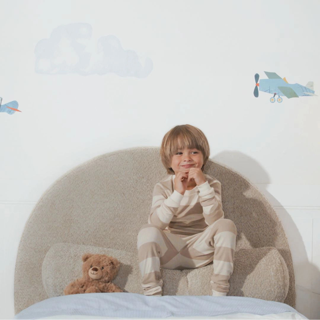 Child in pajamas sitting on a bed with a teddy bear, surrounded by airplane decorations on the wall.