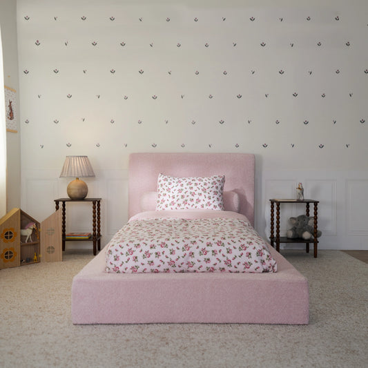 A pink upholstered bed in a child's bedroom, featuring a matching floral-patterned duvet cover and pillow. The room has a white paneled wall decorated with small flower decals, flanked by two dark wood side tables with lamps and toys.
