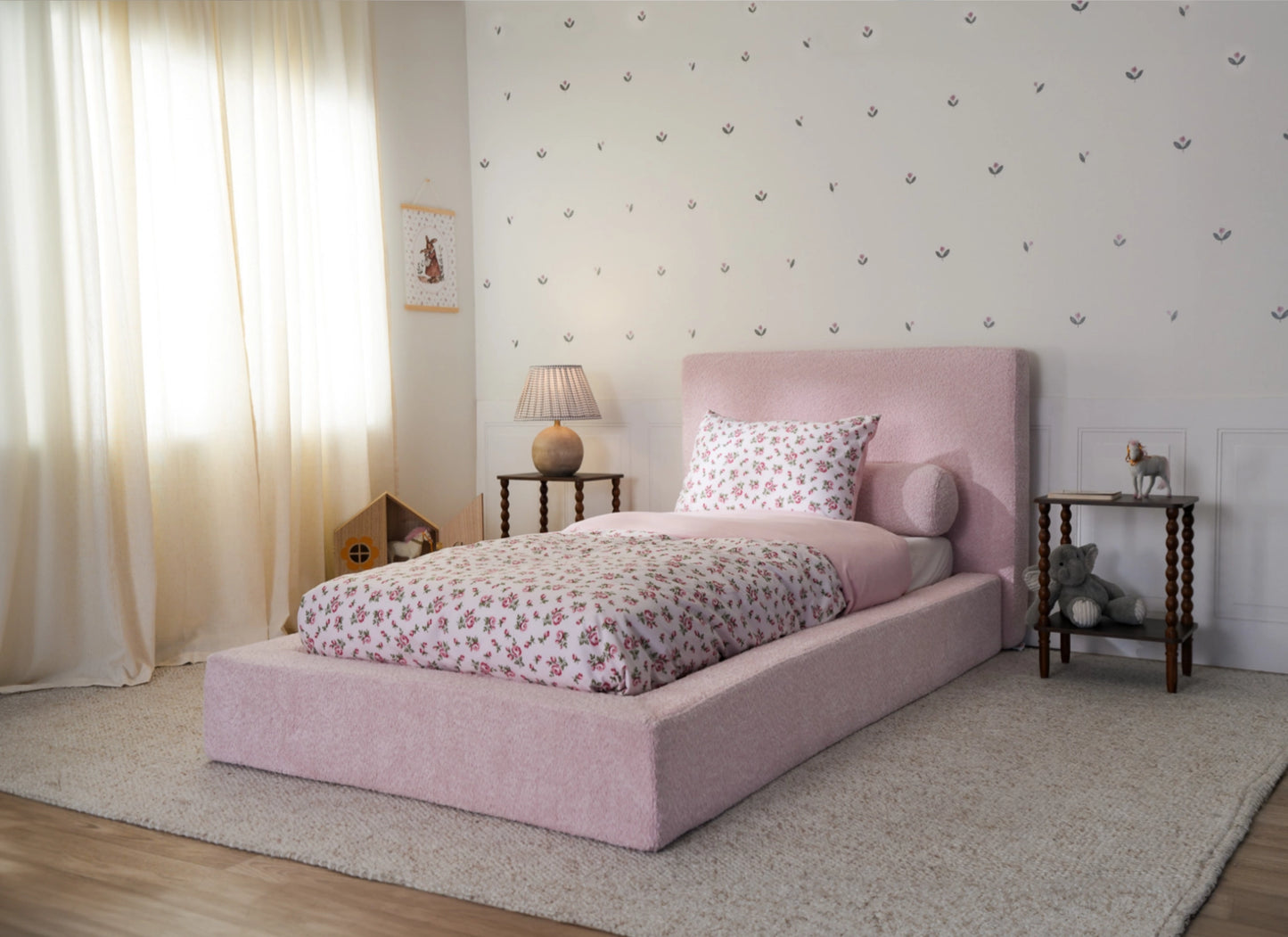 Pink all cushioned bed frame for kids in a twin size with floral sheets and beige curtains.
