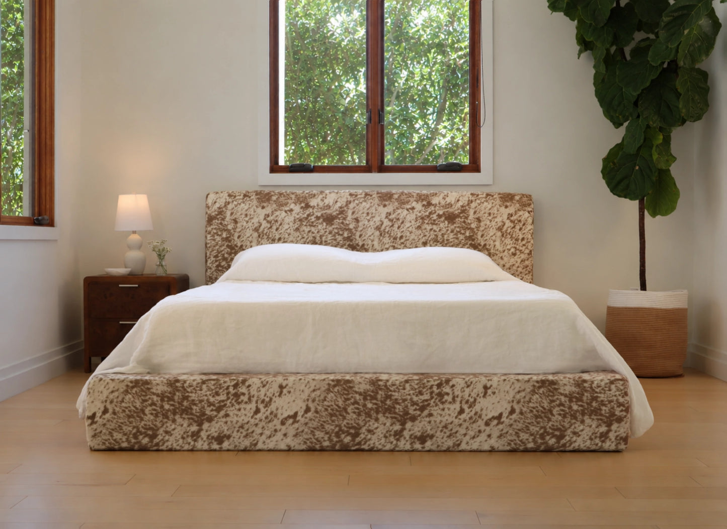 A low-profile platform bed with a textured, mottled brown and beige upholstered frame, dressed in white bedding, set in a bright bedroom with a window, bedside lamp, and potted plant.