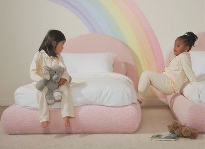 Two children sitting on a pink all cushioned bed with a rainbow mural in the background.