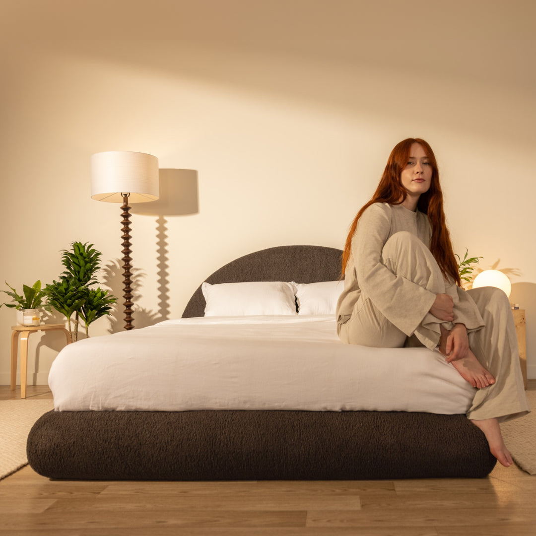 Woman sitting on a dark brown bed in a softly lit bedroom | Luna Set in Chocolate | SoftFrame® Designs