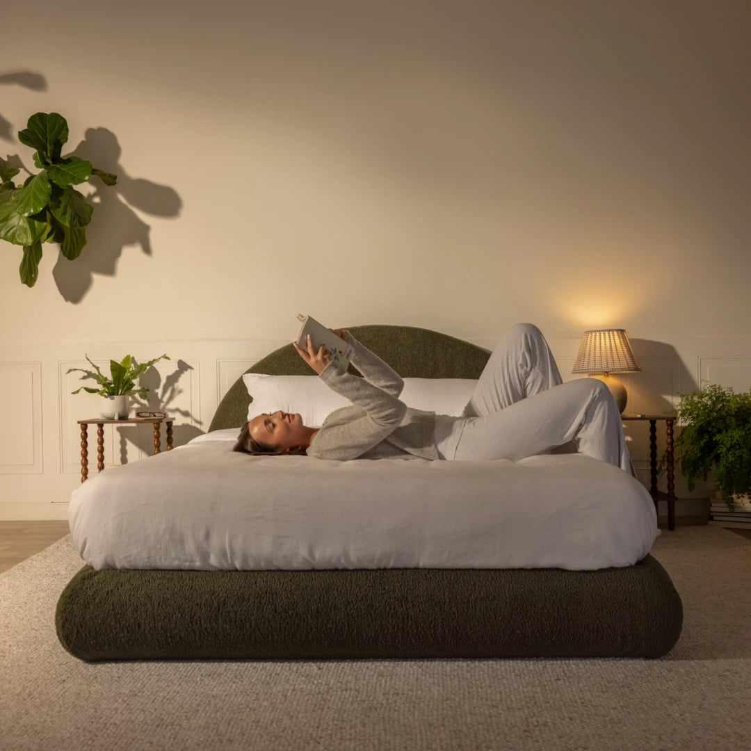 Woman lying on a bed in a cozy bedroom with an olive green, curved bed frame, plants and soft lighting | Luna Set in Moss | SoftFrame® Designs