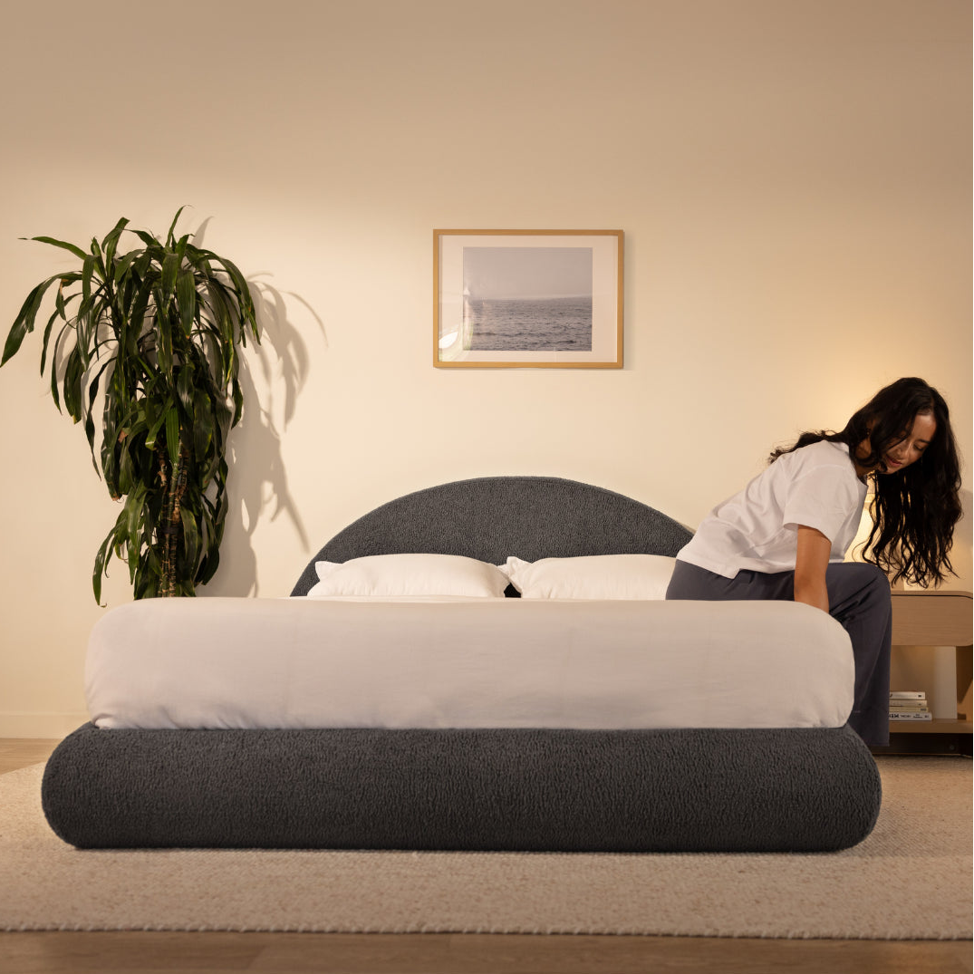 Woman sitting on a queen soft bed in a room with a plant and framed picture on the wall | Luna Set in Storm | SoftFrame® Designs