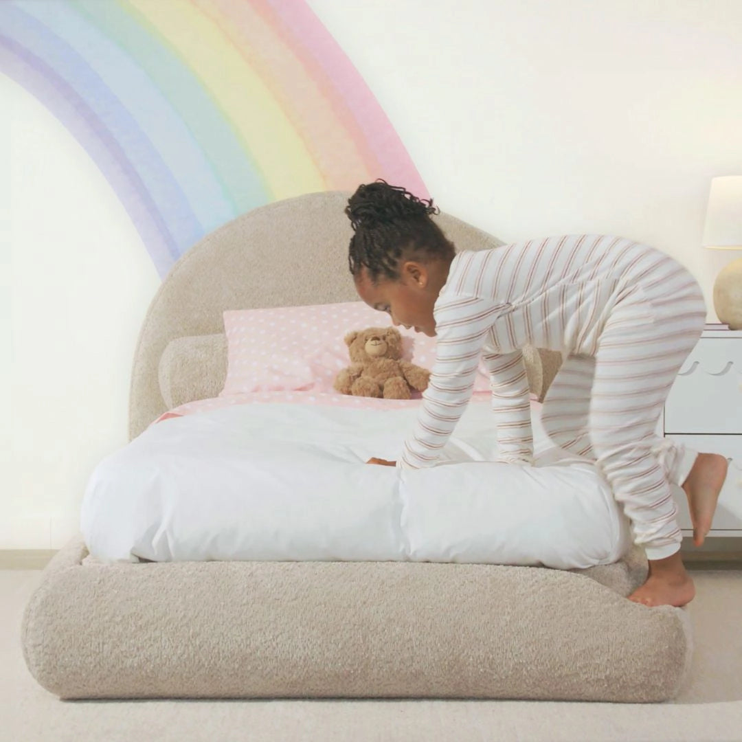 Child sitting on a large bed with teddy bear, airplane wall decals, and nightstand.
