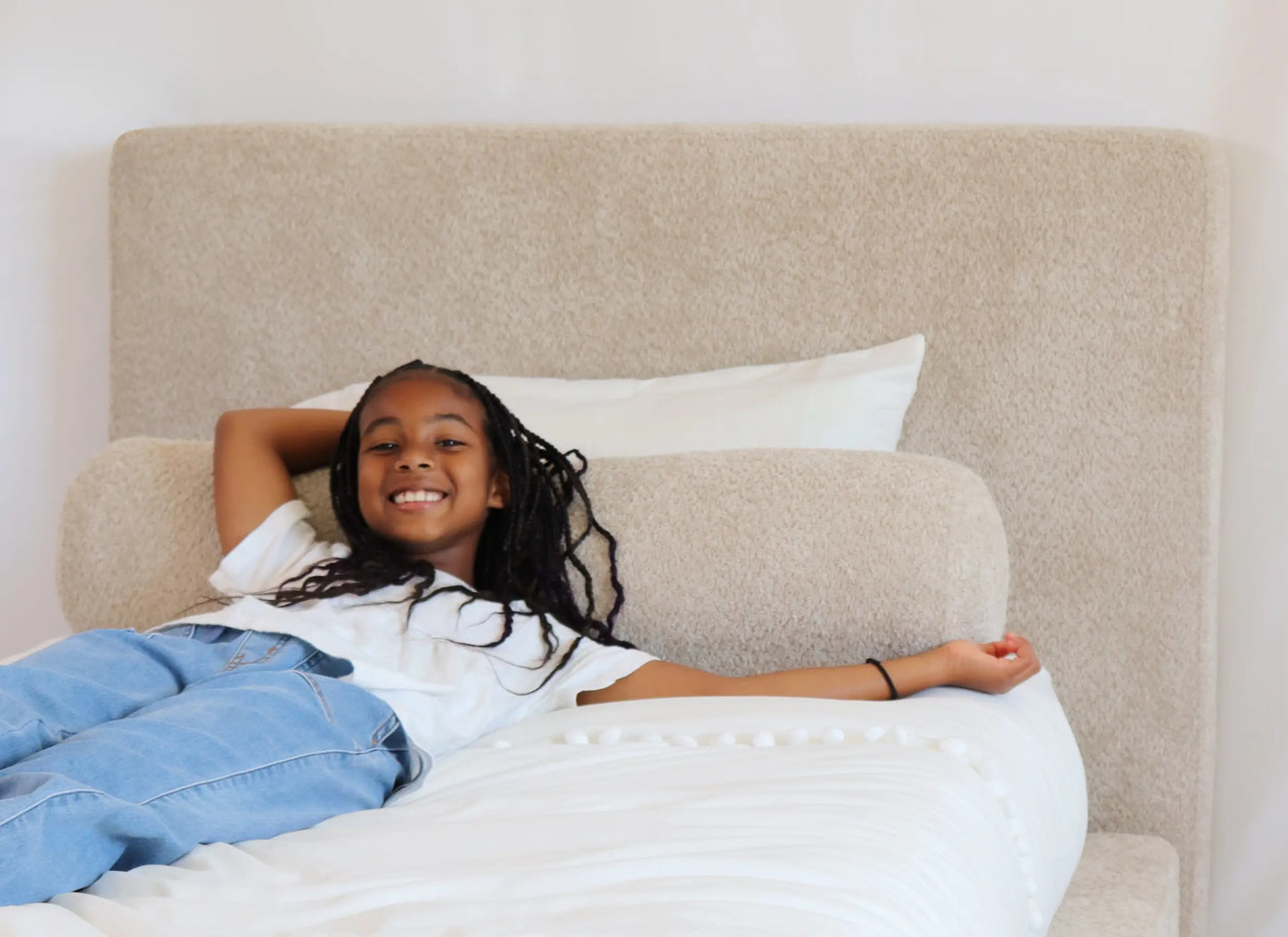 Young girl lying on an all-cushioned bed with a beige all-cushioned headboard and white pillows.
