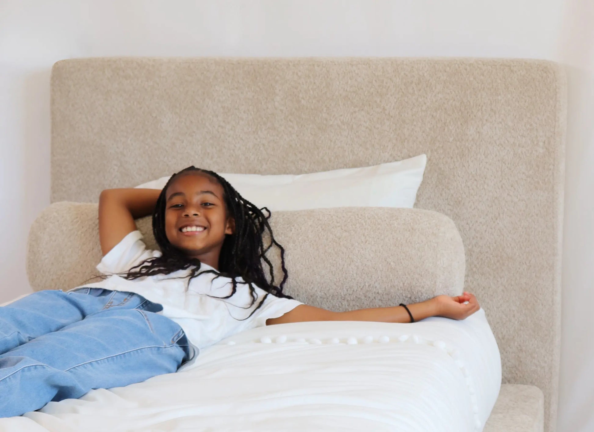 Young girl lying on an all-cushioned bed with a beige all-cushioned headboard and white pillows.
