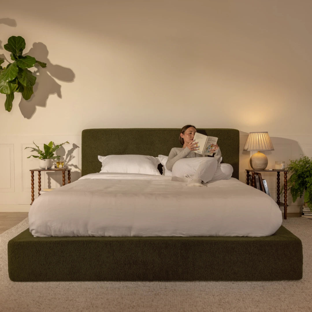 Woman reading a book on a bed in a cozy bedroom setting with plants and lamps | Ultra Set in Moss, olive green | SoftFrame® Designs
