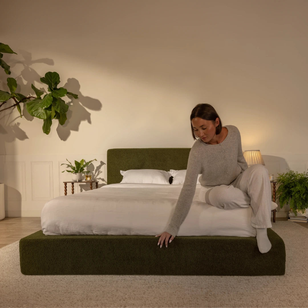 Woman sitting on a bed in a bedroom with plants and a lamp, pressing her hand and foot on a green soft bed | Ultra Set in Moss | SoftFrame® Designs