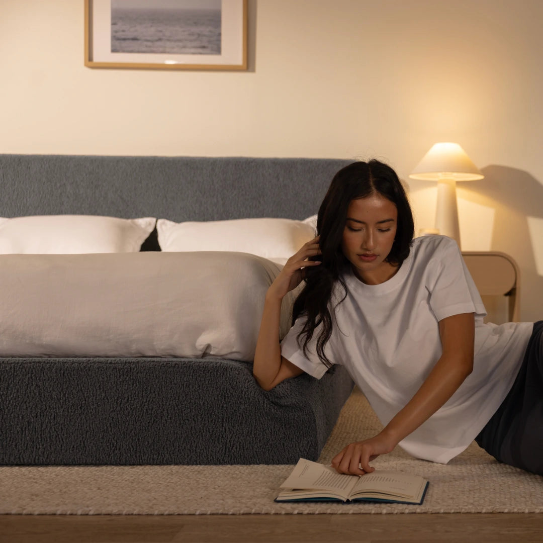Woman reading a book on the floor in a bedroom with a storm grey bed and lamp in the background | Ultra Set in Storm | SoftFrame® Designs