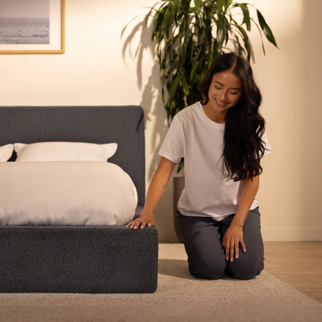 Woman kneeling on the floor next to a soft upholstered, grey bed in a bedroom setting | SoftFrame® Designs