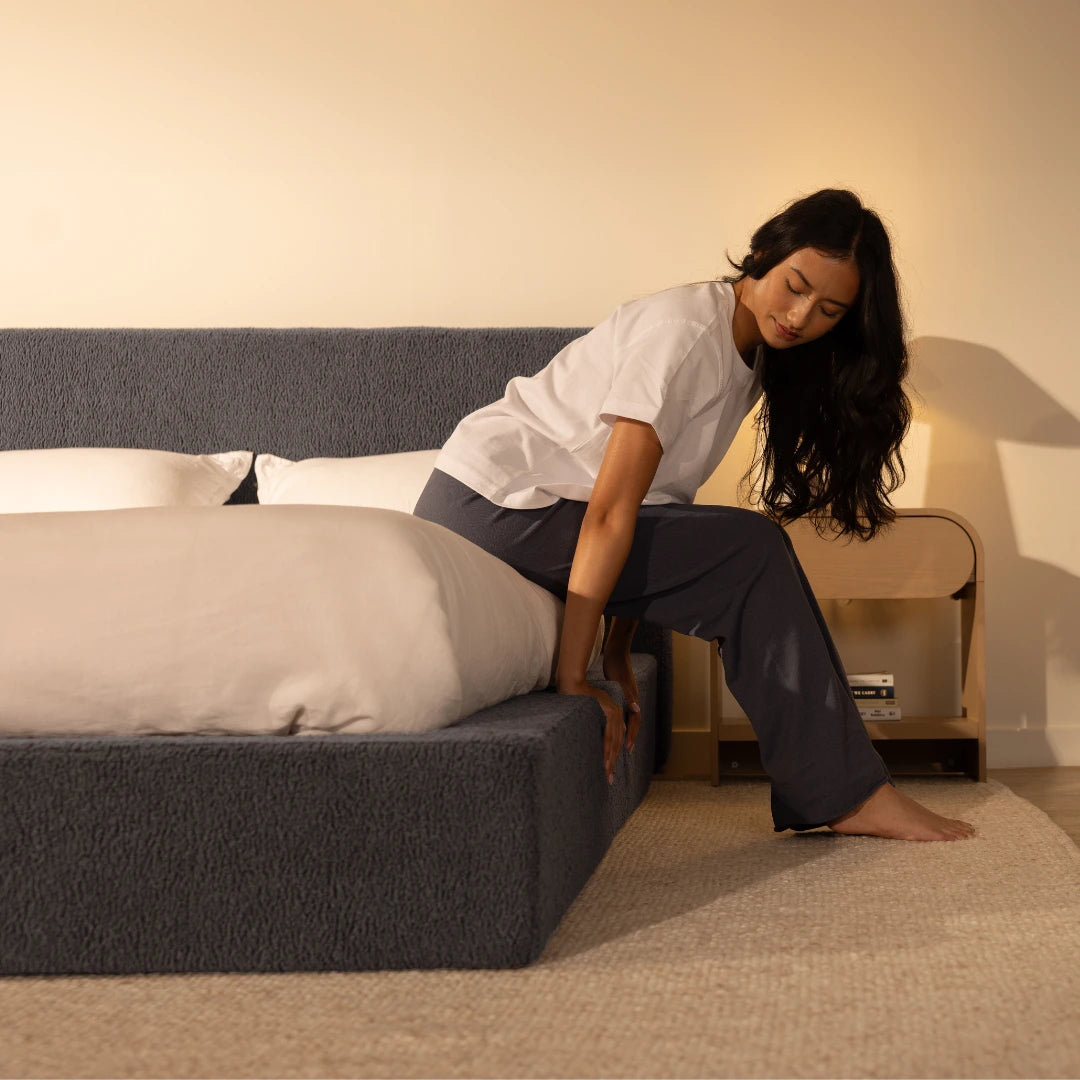 Woman sitting on the edge of a bed and pressing her handing on a soft bed frame in a bedroom setting | Ultra Set in Storm | SoftFrame® Designs