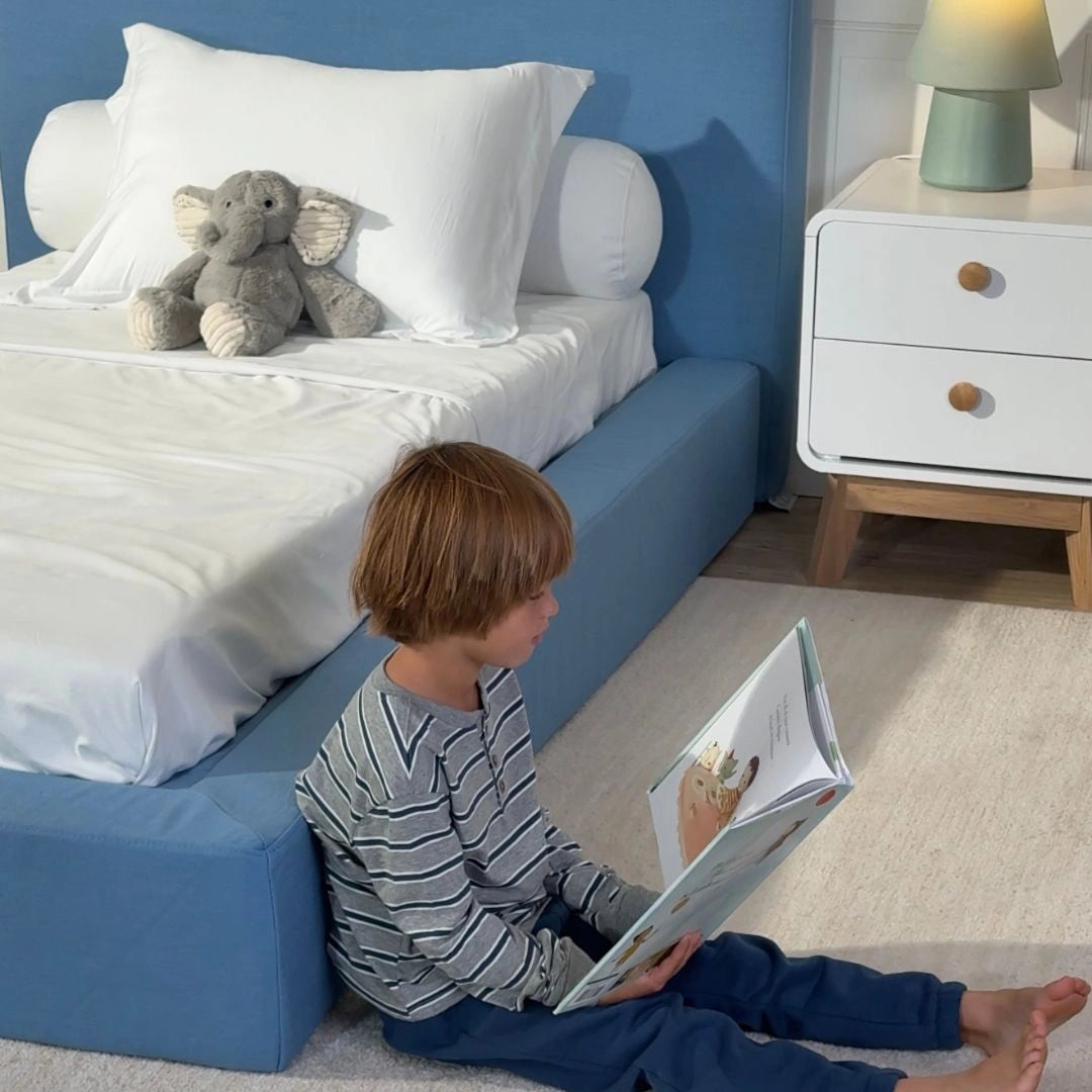 Child reading a book in a bedroom with a blue bed and white nightstand.