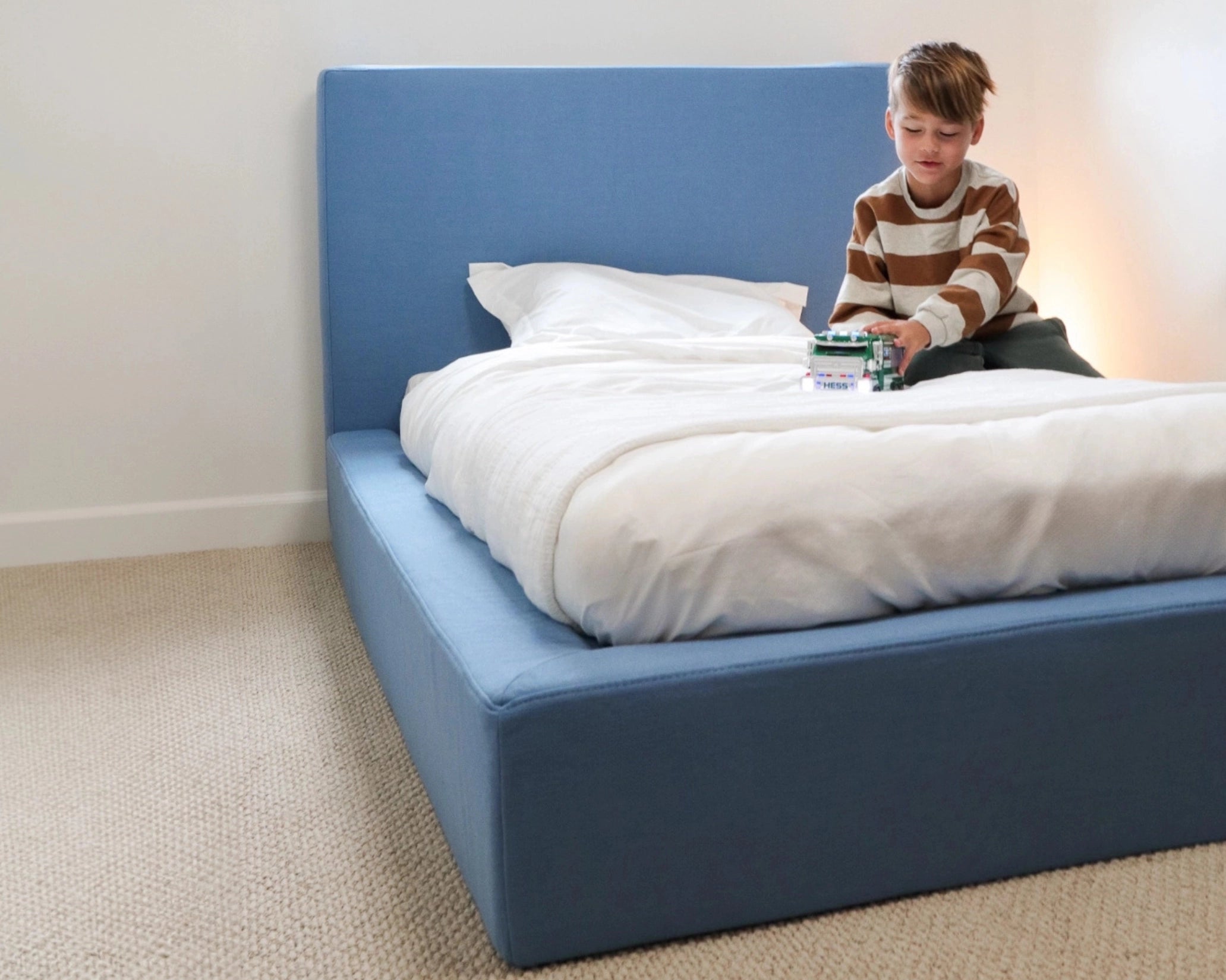 Child sitting on a all cushioned bed holding a teddy bear with a blue all cushioned headboard and airplane-patterned pillow.