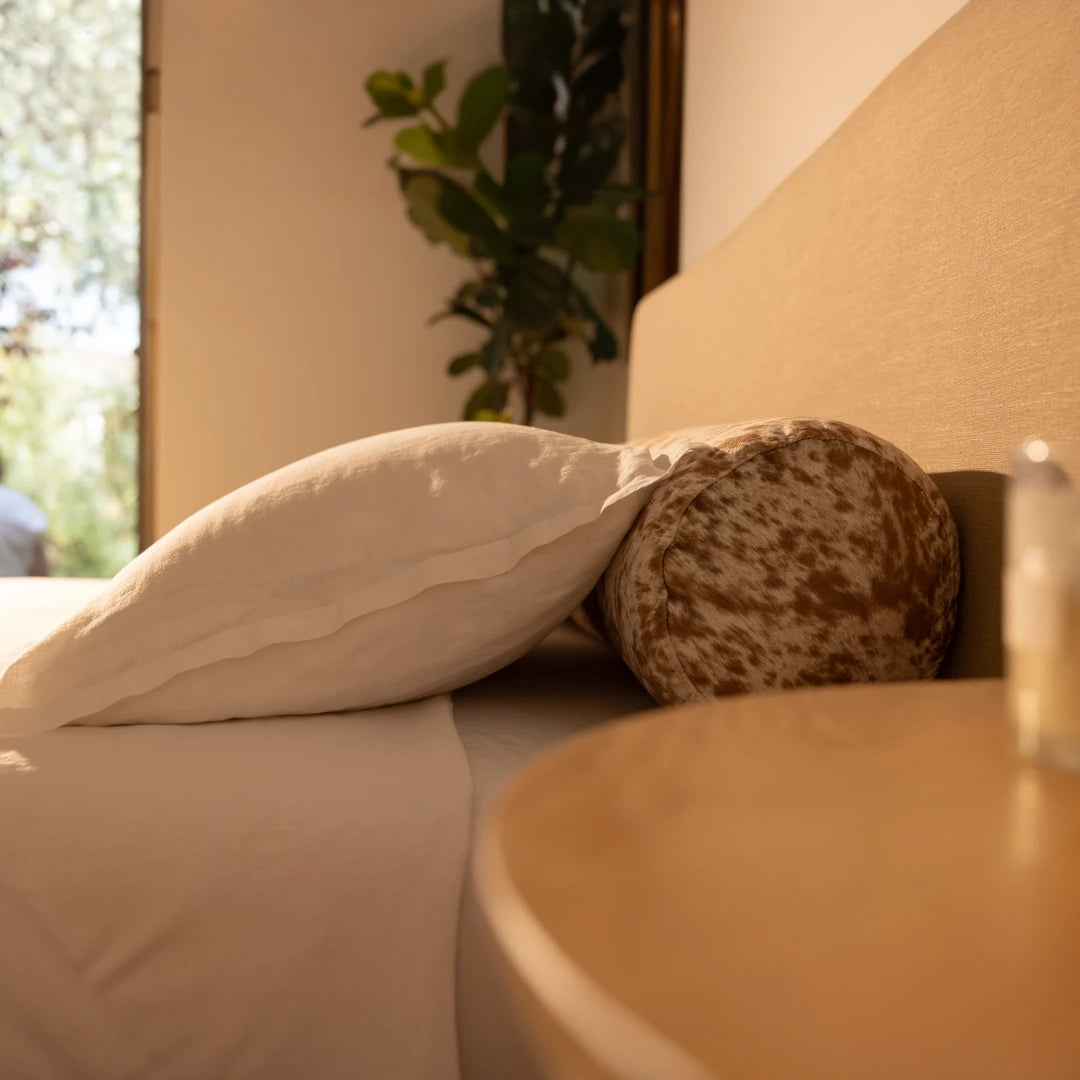 Close-up side view of a bed with pillows, a round bolster pillow, and plant in the background | Round Bolster Pillow, Queen, Pony | SoftFrame Designs