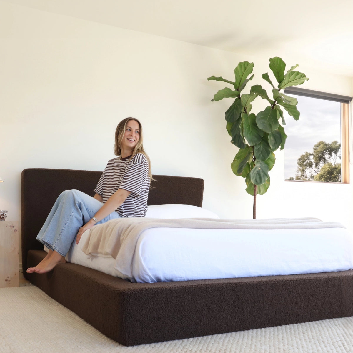 Ultra all cushioned bed frame and headboard in the fabric Chocolate in a bright minimalist room with a woman sitting on th bed