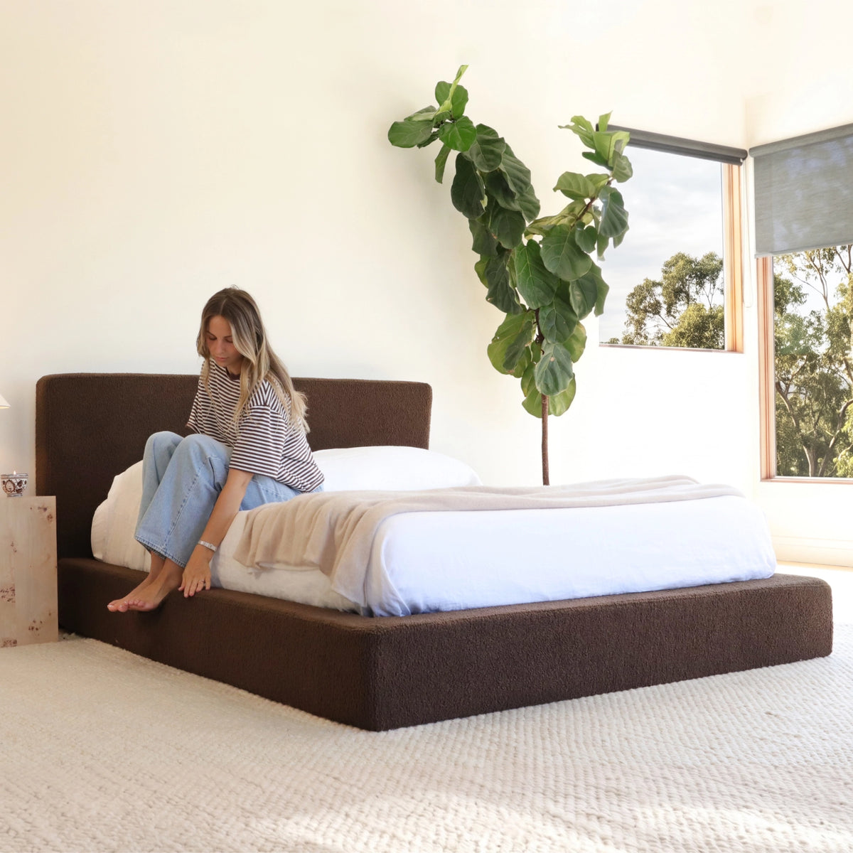 Ultra all cushioned bed frame and headboard in the fabric Chocolate in a bright minimalist room with a woman sitting on th bed