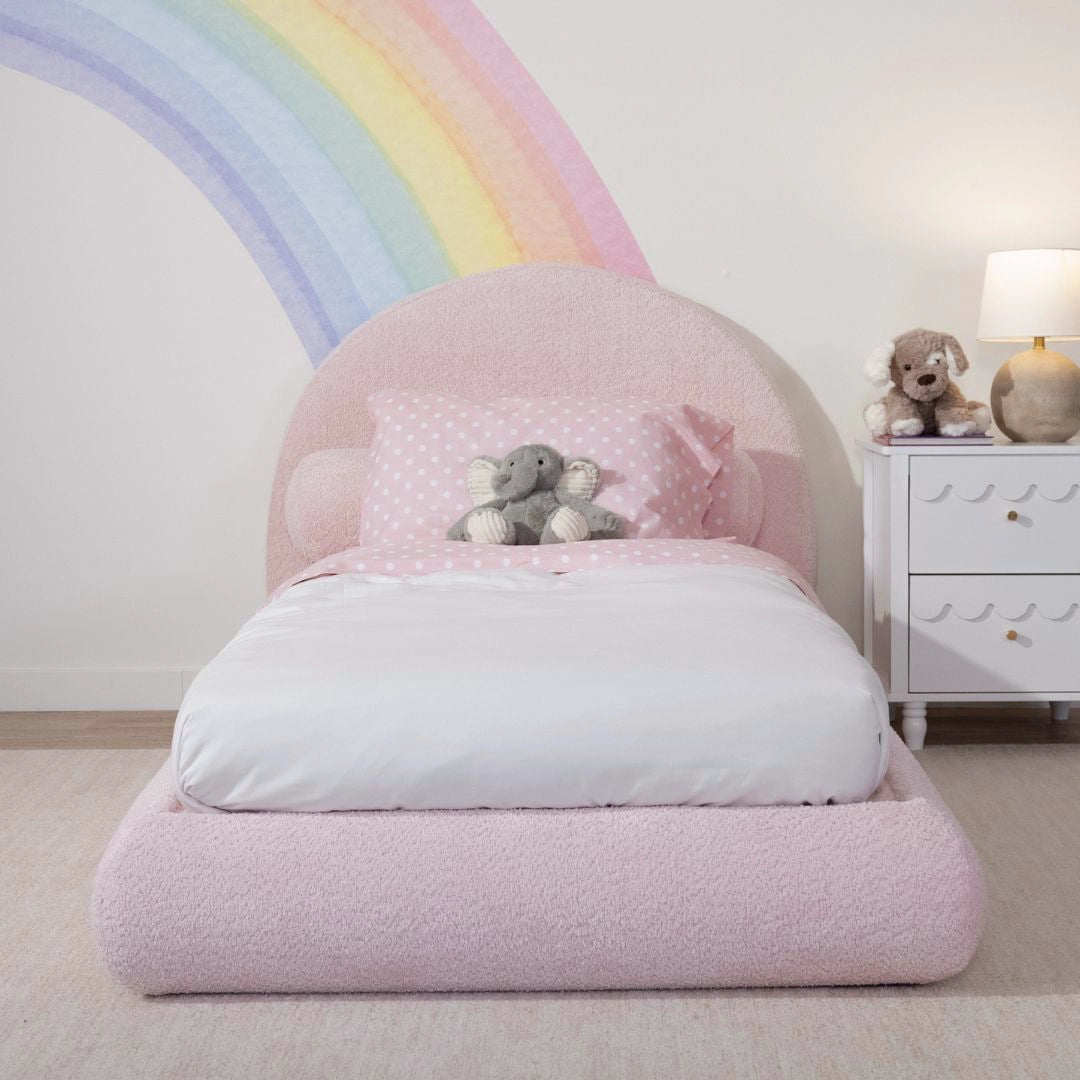 Pink children's bed with a rainbow on the wall and a teddy bear on it, in a room setting.