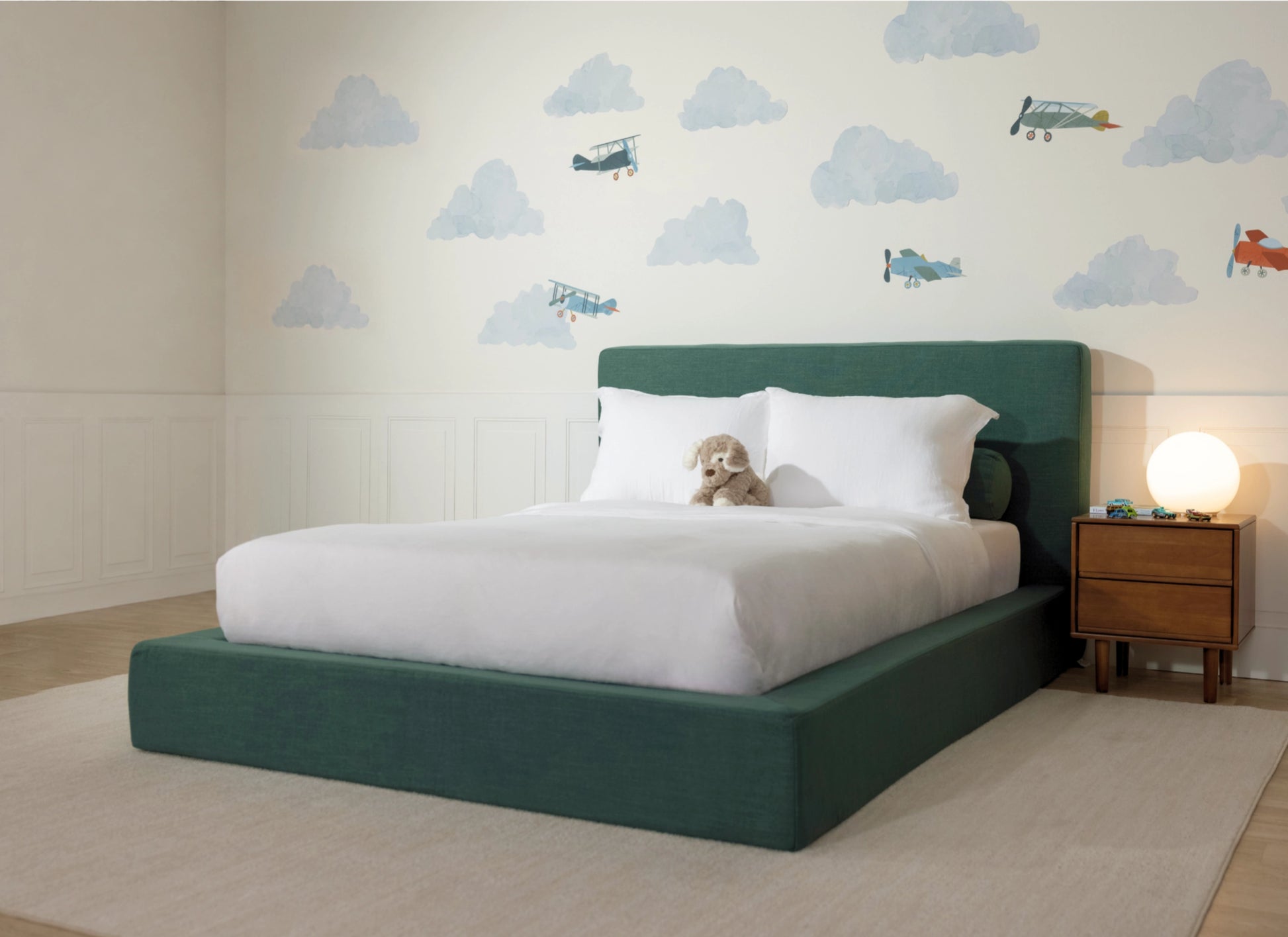 A modern platform bed with a dark green upholstered frame and headboard, dressed in white bedding, set in a child's bedroom with cloud and airplane wall decals.