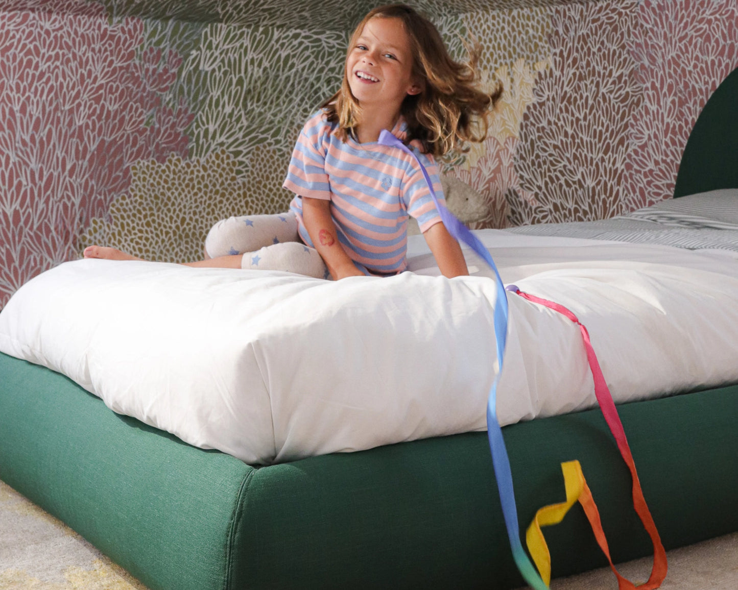 Child sitting on a all cushioned bed frame with colorful ribbons, smiling.