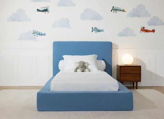 A modern children's bedroom featuring a low-profile platform bed with a light blue upholstered headboard and footboard, dressed in white bedding with a small gray stuffed animal on the mattress. The room has a white paneled wall decorated with blue and red vintage airplane wall decals, a wooden nightstand with a glowing globe lamp, and a light beige rug.