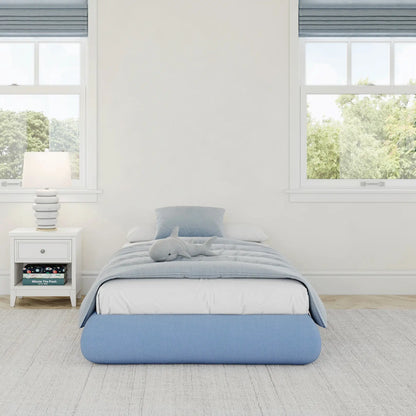 A front view of a kids all cushioned bed frame in sky in a gray kids bed room with big windows with trees. 