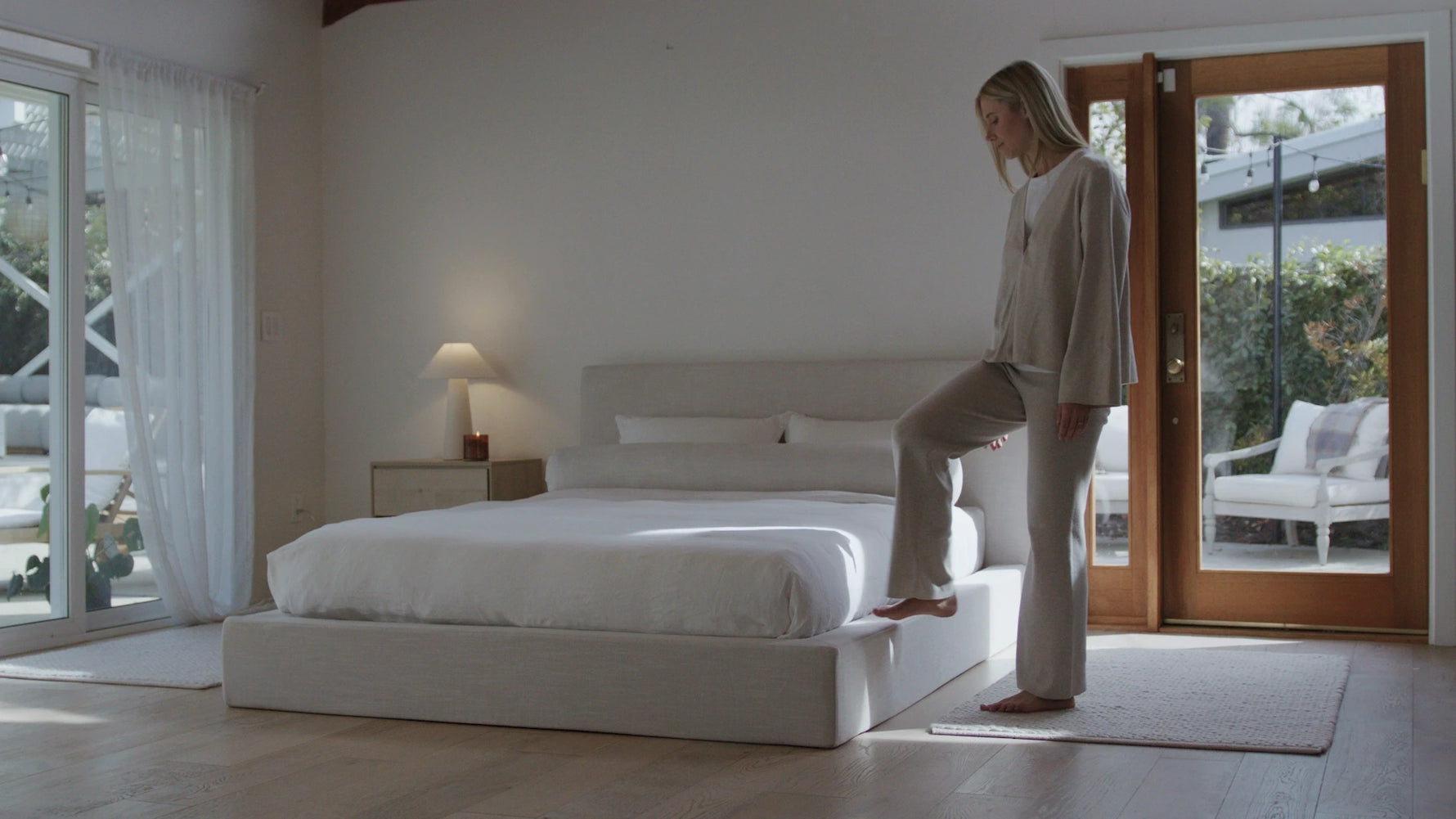 Woman standing on a white platform bed in a modern bedroom with large windows.
