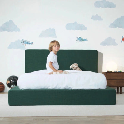 Child sitting on a green bed with a white mattress, surrounded by cloud-patterned wallpaper.