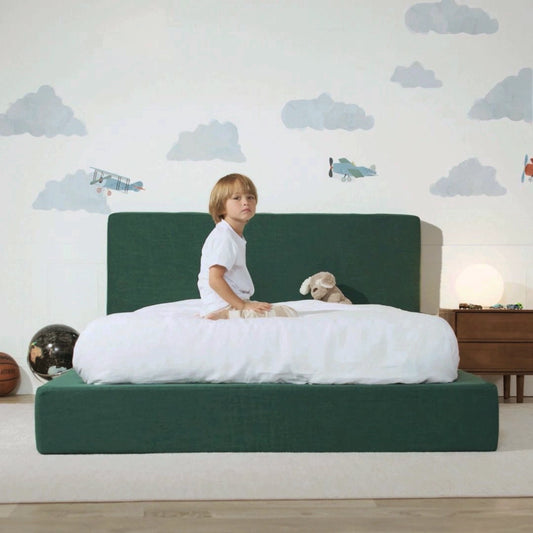 A child sits on a low-profile platform bed with a dark green upholstered headboard and footboard, dressed in a white t-shirt and white bedding. The room features a light wall decorated with gray cloud decals and small airplane illustrations, a wooden nightstand with a glowing lamp, and a small globe on the floor.