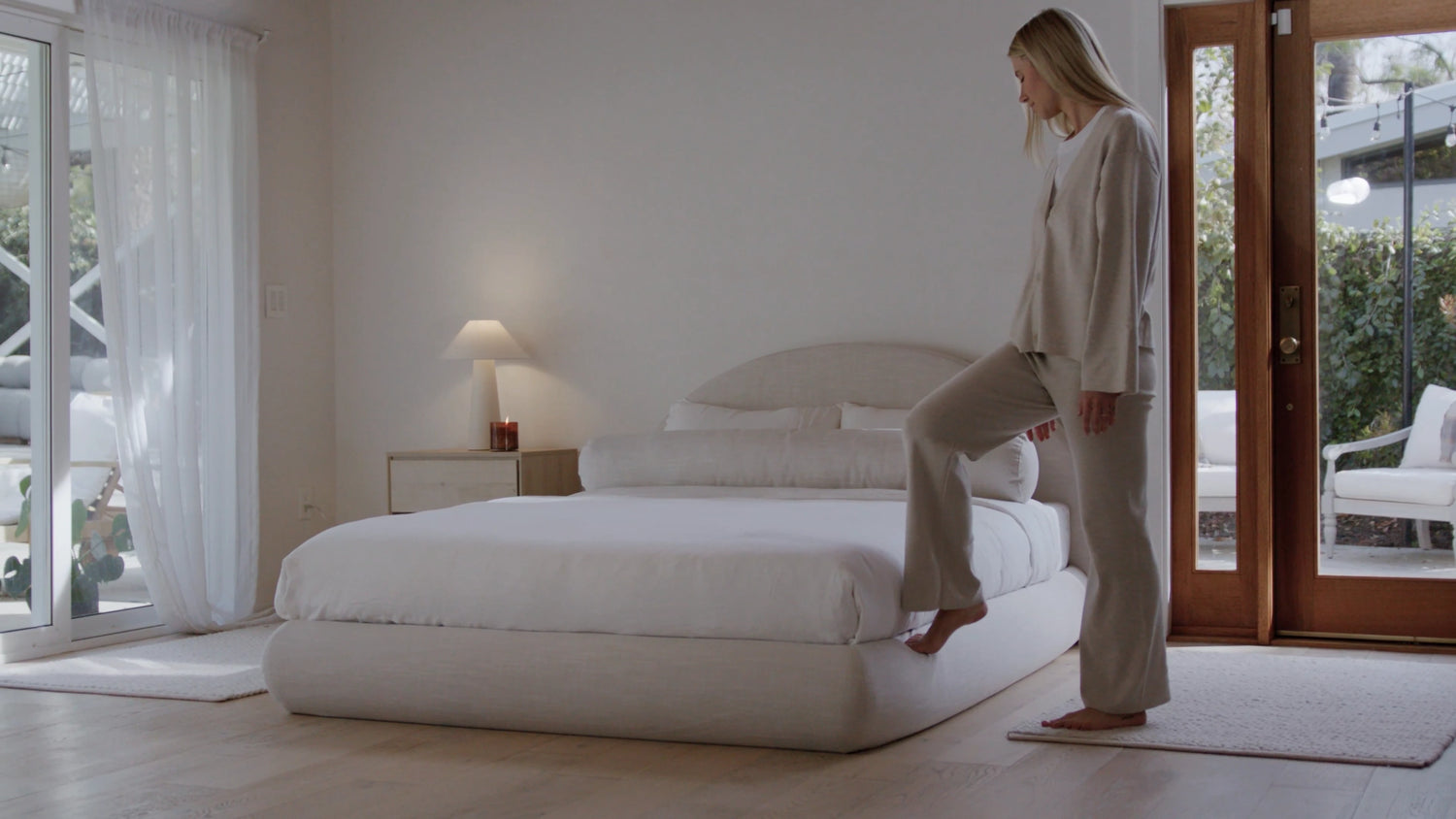 Woman standing by a soft, curved bed in a modern bedroom with large windows.