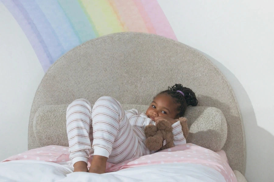 Child lying on a bed with a teddy bear, rainbow decoration in the background | SoftFrame Kids