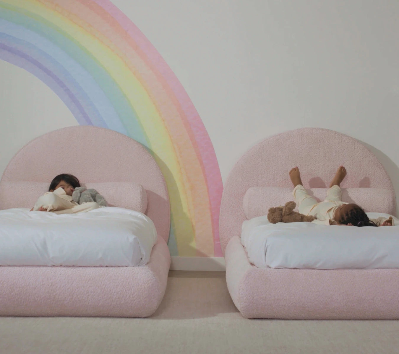 Two children lying on pink beds with a rainbow mural in the background | SoftFrame Kids