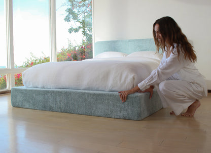 Woman in a white outfit adjusting a bed in a bright room with large windows.