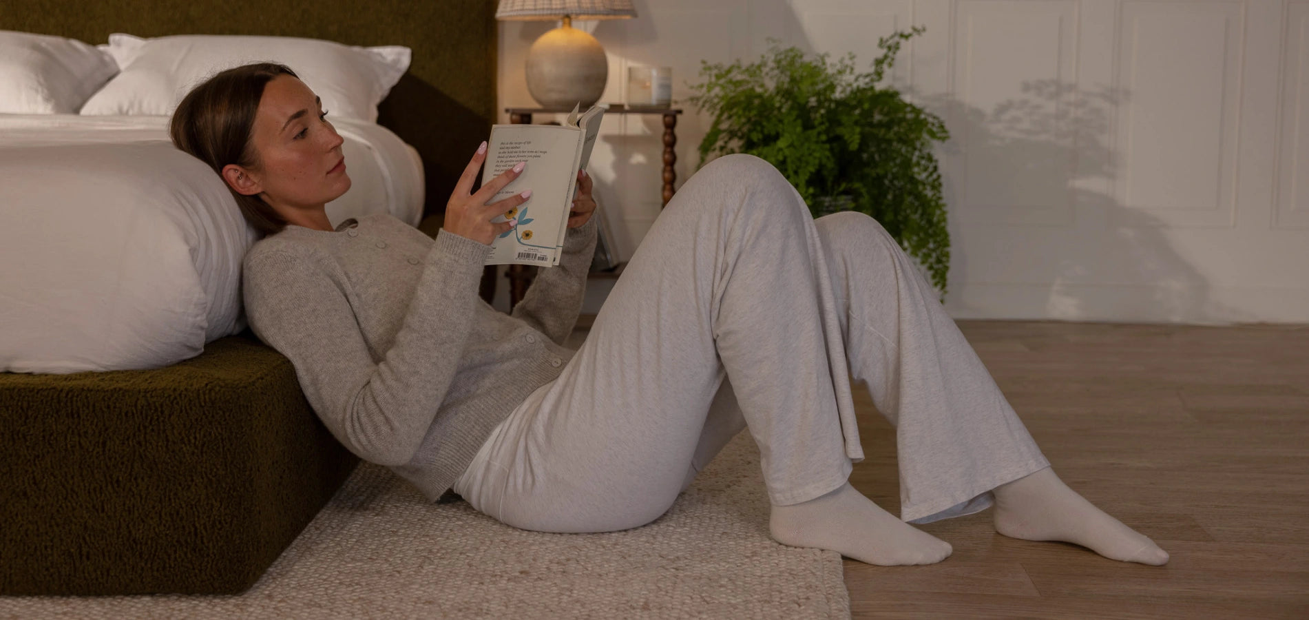 Woman reading a book on the floor while leaning on bed in a cozy bedroom setting