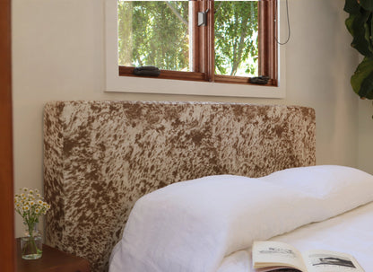 A close-up of a bed with white linens and a tall upholstered headboard in a brown and cream mottled pattern, set in a bedroom with a window and a small vase of flowers on a bedside table.